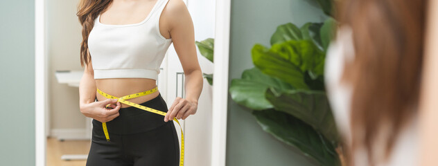 Weight lose, loss concept. Slim body asian young woman hand use tape measuring around waistline in fit sports. Girl looking reflect in mirror at home. Healthy nutrition, fitness for wellbeing beauty. - obrazy, fototapety, plakaty