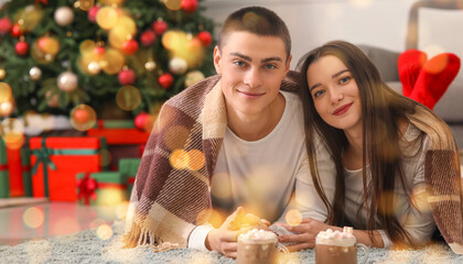 Young couple with plaid and cups of hot chocolate lying at home on Christmas eve