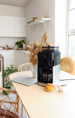 Modern coffee machine with cup, bun and laptop on table in kitchen