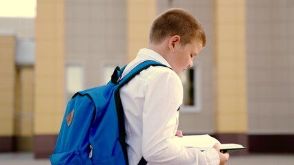 Pupil child hurries to school for lesson and reads book on city street. Child with book and backpack hurries to school. Boy kid walks with textbook in his hand. Concept of children education, learning