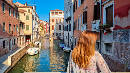Rear view of woman watching water channel in city Venice, Veneto, Italy, Europe. Venetian architectural landmarks and old houses facades along. Urban tourism in summer atmosphere. Romantic vacation - obrazy, fototapety, plakaty