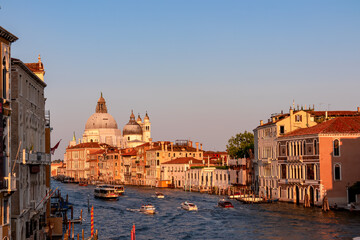 Panoramic sunset view of Grand Canal in city of Venice, Veneto, Italy, Europe. Famous landmark cathedral Santa Maria Della Salute along the water traffic corridor Grand Canale. Urban tourism in summer - obrazy, fototapety, plakaty
