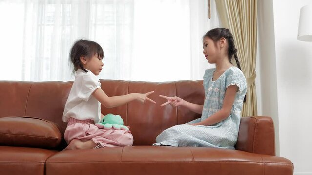Two little Asian girls play with rocks, paper, scissors on the sofa in their home. Both of them are happy and having fun. Young children in elementary