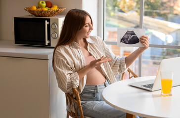 Young pregnant woman with sonogram image video chatting at table in kitchen