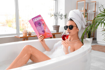 Beautiful woman in sunglasses with magazine and wine taking bath at home