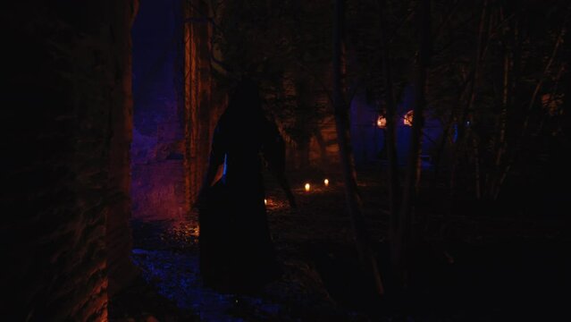  In a following shot, a gorgeous witch woman walks around an abandoned church with candles around, playing with her black dress. The scene takes place outdoors during the night.