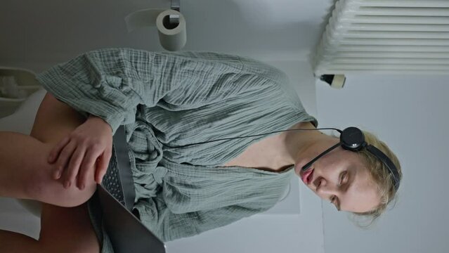 A woman programmer sits on the toilet with headphones and a laptop, talks with colleagues on Skype, participates in an online conference. Vertical video