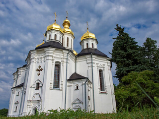 The Orthodox Church of St. Catherine in Chernihiv, with its white facade, iconic golden domes, and crosses, harmonizing beautifully with the vibrant greenery and the serene blue sky.