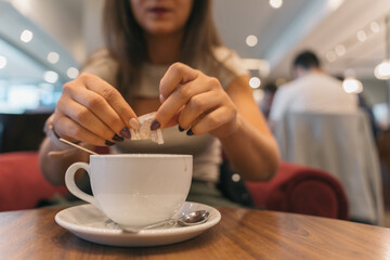 Unrecognizable woman pouring a packet of sweetener into her coffee. Young anonymous girl sweetening her freshly brewed coffee at the airport restaurant while connecting flights.