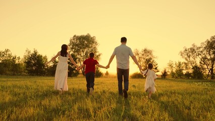 Happy family run forward on green grass park. Mom, dad, daughter, son holding hands running across field. Parents children hold hands run towards sunset. Mother father kid play in nature. Young family - Powered by Adobe