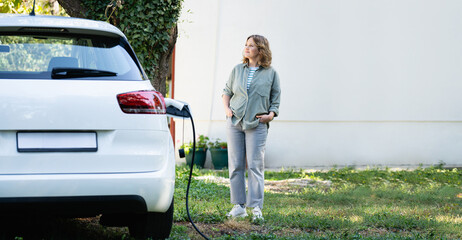 Woman next to a charging electric car in the yard of a country house