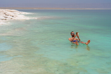 Relaxing tourist man reading a book while taking a bath in floating Death Sea, Jordan