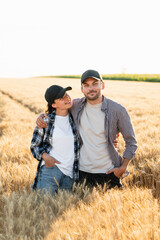 Naklejka na ściany i meble A couple of farmers in plaid shirts and caps stand embracing on agricultural field of wheat at sunset.