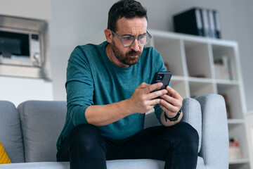 Worried mature man thinking about what he has read on his mobile phone while sitting on the couch...