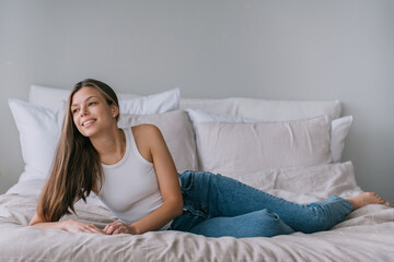 Obraz na płótnie Canvas Relaxed young woman lounging on a bed, casually dressed in a white tank top and blue jeans, with a serene smile, embodying comfort and effortless style in a cozy setting.
