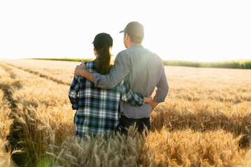 A couple of farmers in plaid shirts and caps stand embracing on agricultural field of wheat at...