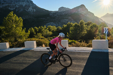 Fit woman cyclist wearing cycling kit and helmet riding on the road on a gravel bike at sunset. Empty mountain road. Sports motivation image. Bernia mountain in Spain. - obrazy, fototapety, plakaty