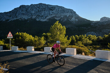 Woman cyclist wearing cycling kit and helmet riding on the road on a gravel bike at sunset.Empty mountain road. Sports motivation image. Bernia mountain in Spain.