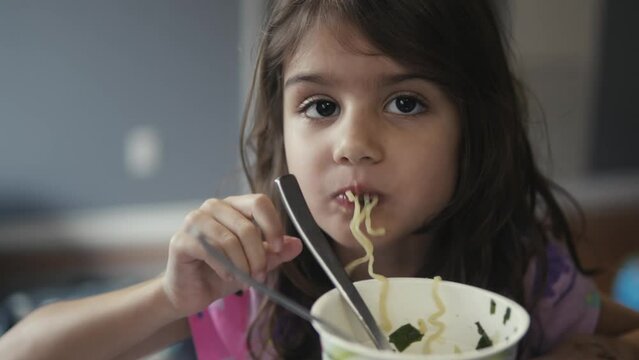 Close-up of a 5-year-old girl delightfully eating ramen noodles with a fork, showcasing the simplicity and joy of childhood meals