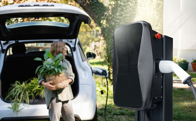 Woman with plant in pot next to a charging station and electric car in the yard of a country house