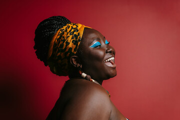 Happy plus size African woman with colorful make-up wearing traditional headwear on red background - obrazy, fototapety, plakaty
