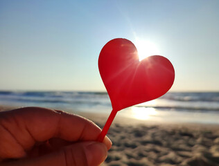 Person holding in fingers hand stick in shape red heart on background sea and sea waves, blue sky in seashore on sunny summer day close-up. Concept love romance amour St Valentines Day - obrazy, fototapety, plakaty