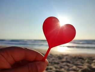 Fotobehang Person holding in fingers hand stick in shape red heart on background sea and sea waves, blue sky in seashore on sunny summer day close-up. Concept love romance amour St Valentines Day © mari1408