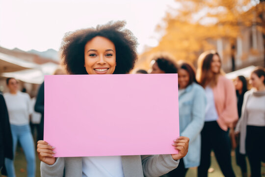 A young African American woman smiling and holding a blank rectangular pink sign to add text on it, claiming, women in the background blurred at a demonstration.Mockup