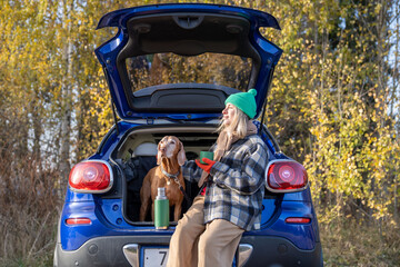 Woman pet owner drinking tea sitting in car trunk with dog resting on nature in forest on weekend....
