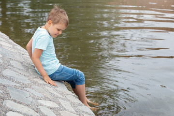 A happy boy is sitting on a stony pond bank in a city park and soaking his feet in the water