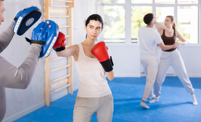 Active young girl training boxing kicks on punch mitts held by instructor in sports hall