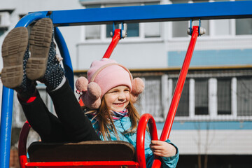 A funny, smiling girl, a preschool child rides, sitting on a metal red swing, on the playground....