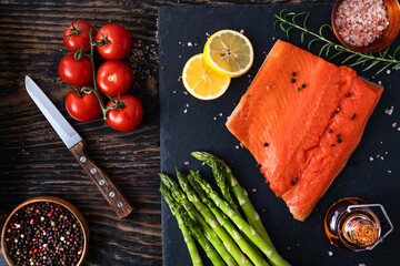 salmon fillet with salt and pepper on a stone slab. Asparagus and lemon on dark wooden table. View...