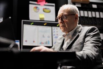 Old business manager executive sitting at office desk before consulting meeting, inspecting quarterly company results analysis data paperwork on laptop. Bureaucratic workplace filled with flowcharts