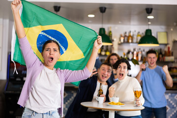 Company of emotional young adult fans supporting sports team of Brazil with state flag while...