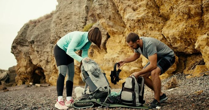 A blonde girl in a blue jacket and a brunette guy in a gray T-shirt lay out and take out ammunition from their backpacks on a rocky beach near the yellow rocks, they will be rock climbing during the