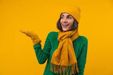 Portrait of attractive smiling young woman posing in cozy knitted sweater, beanie hat and scarf, isolated over yellow background