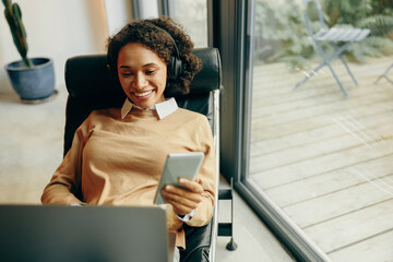 Smiling female freelancer working on laptop from home while use mobile phone sitting in chair
