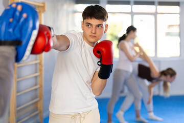 Sportive young male practitioner of boxing courses applying kicks on punch mitts during workout...