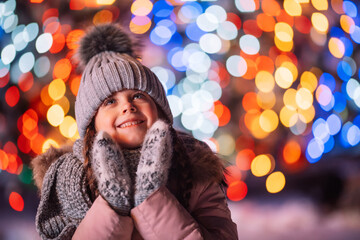 On Christmas night a little girl looks up at the sky outdoors on blurry lights. There is frost and snow outside. City streets illuminated for Christmas