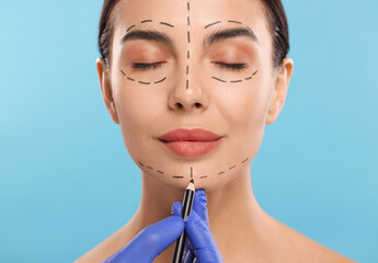 Doctor drawing marks on woman's face for cosmetic surgery operation against light blue background