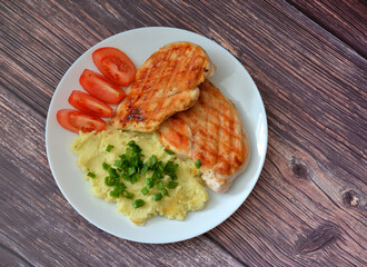 Two grilled chicken fillets with tomatoes and mashed potatoes with green onions on a wooden table.