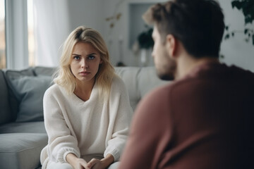 Оffended young blonde woman and man argument, сlose up husband in front and upset wife behind sit at sofa quarrel at home. Family conflict, crisis, psychological abuse, relationships concept - obrazy, fototapety, plakaty