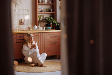 Fototapeta na wymiar A young blonde woman in dark white home clothes sits on a wooden kitchen and drinks hot chocolate with marshmallows. Christmas atmosphere.