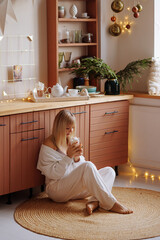 A young blonde woman in dark white home clothes sits on a wooden kitchen and drinks hot chocolate with marshmallows. Christmas atmosphere.