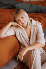Obraz premium A young blonde woman wearing casual clothes lies with a gift in her hands on a bed with an ocher bedspread in a bedroom decorated for Christmas. Cozy design and natural wooden furniture.