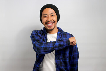 Smiling, happy young Asian man, dressed in a beanie hat and casual shirt, confidently gives a fist...