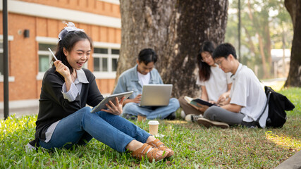 A positive Asian female college student is using her tablet while sitting with her friends in a park