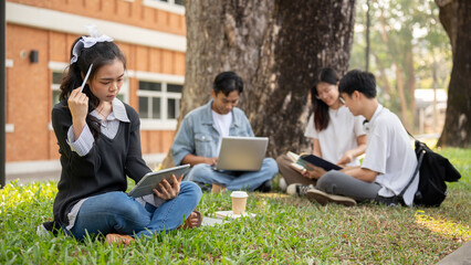A focused Asian female college student is using her tablet while sitting in a park with her friends