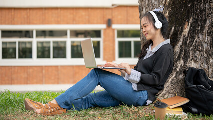 Fototapeta premium A positive young Asian female college student is using her laptop computer under the tree in a park.